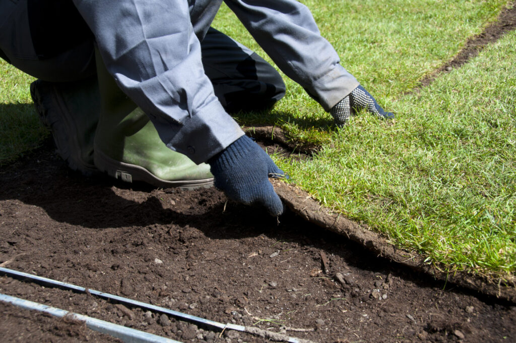 A person in grey pants and green boots is laying sod on soil, with hands visible adjusting a strip of grass beside a metal guide wire.