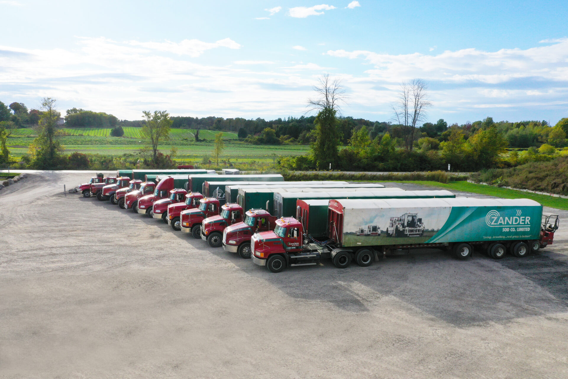 A fleet of red trucks is parked in a line on a gravel lot, with one truck pulling a large green and white trailer.