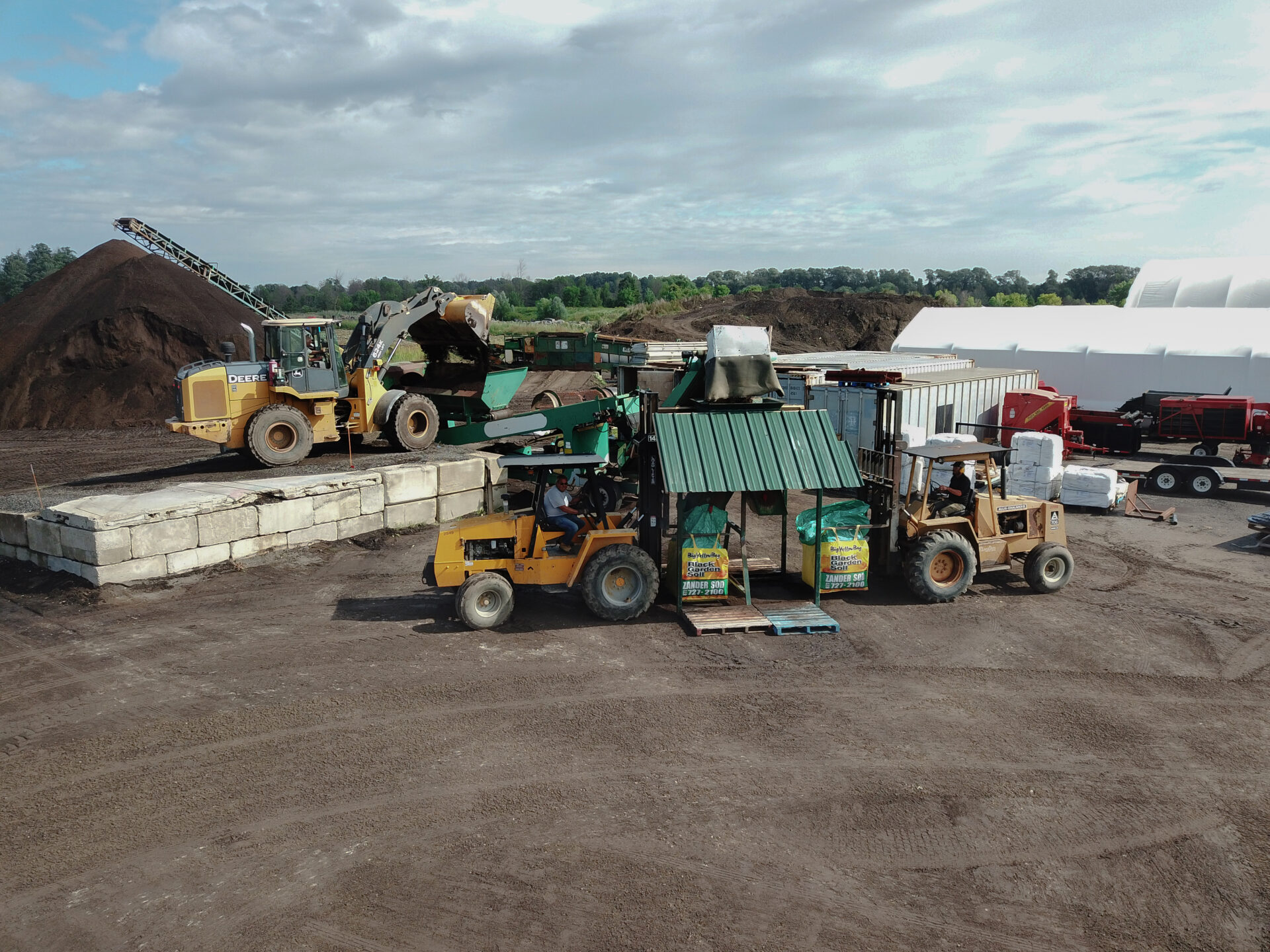 An industrial site with heavy machinery: loaders moving organic material, packaged goods on pallets, and a large compost pile in the background.