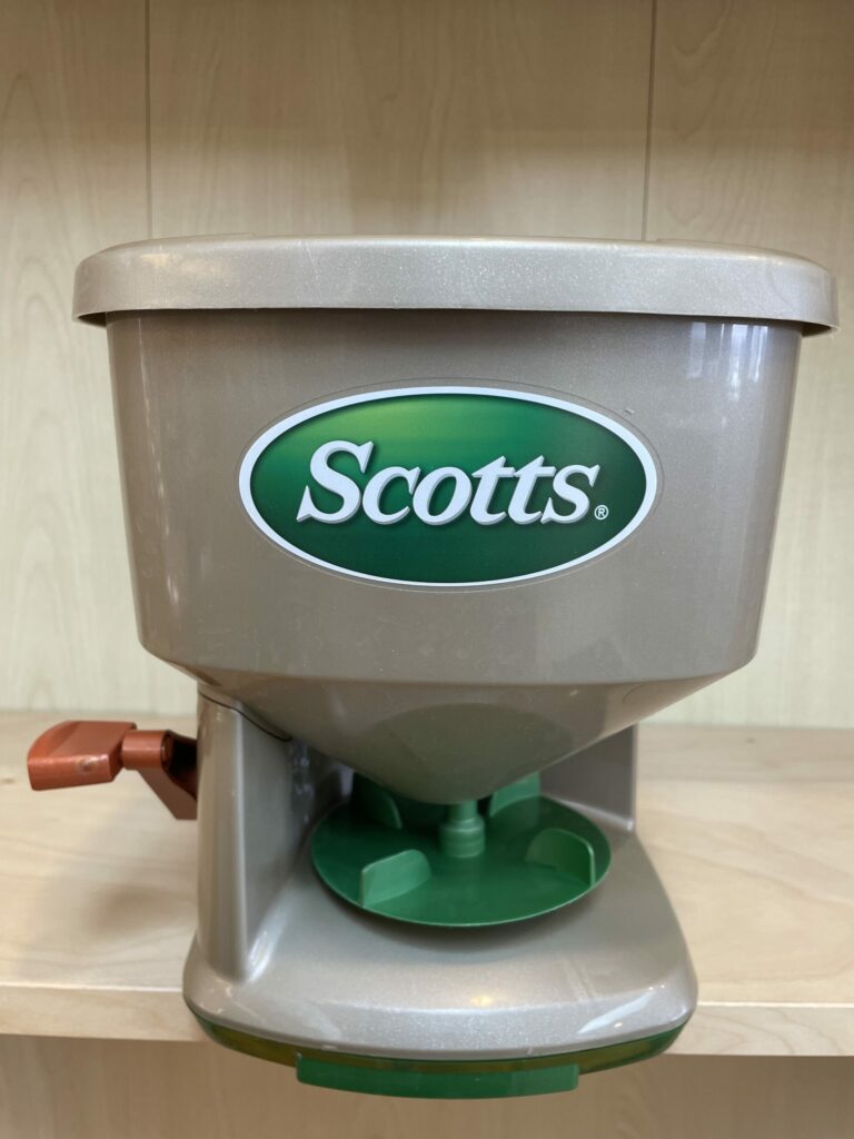 This is an image of a Scotts brand broadcast spreader with a beige hopper, green base, and a brown handle, used for dispersing seeds or fertilizer.