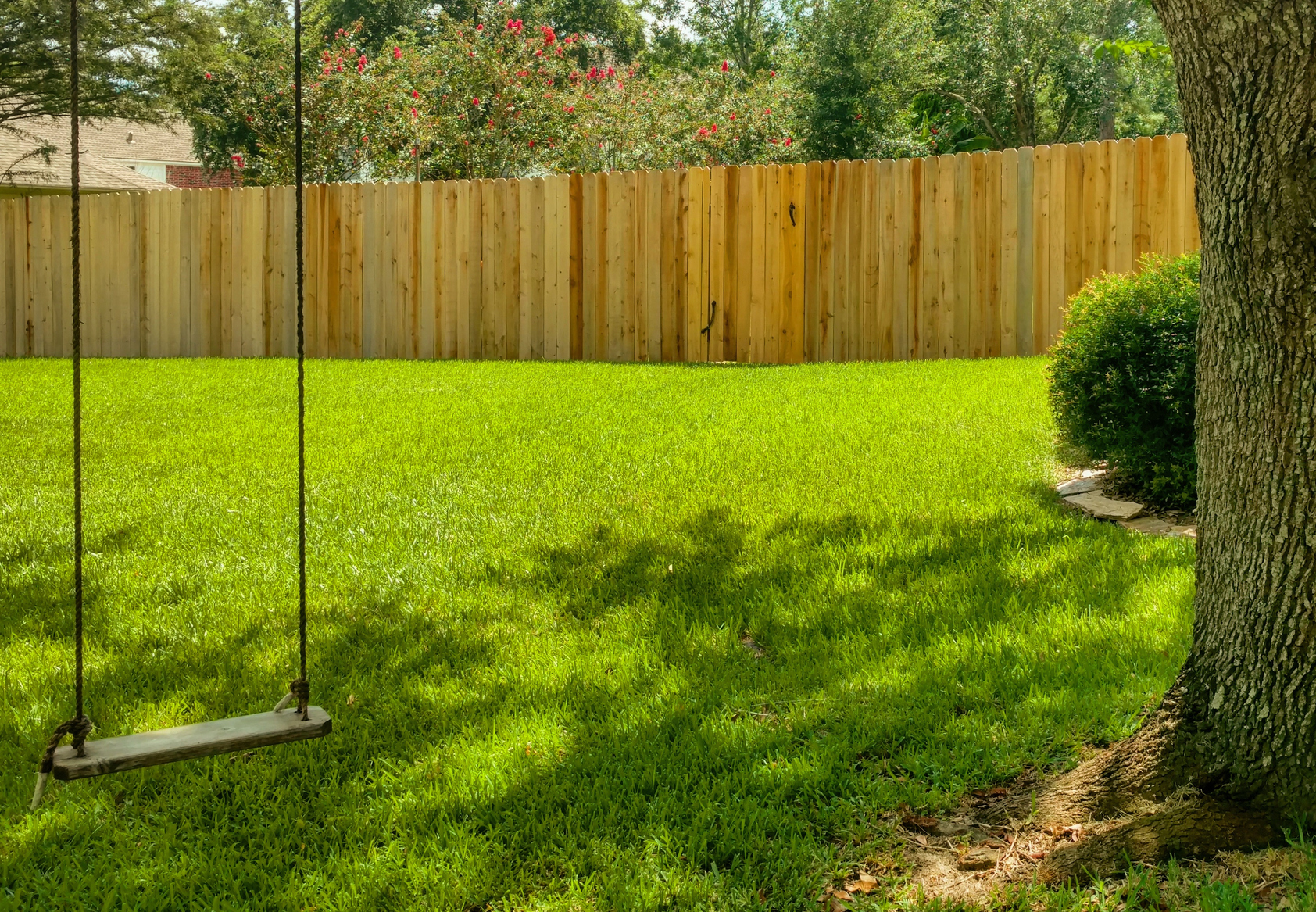 A serene backyard with a lush green lawn featuring a wooden swing hanging from a tree, bordered by a tall, solid wooden fence.