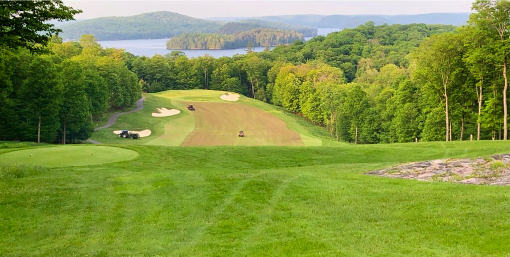 Lush golf course with green fairways, sand bunkers, and golf carts, overlooking a tranquil lake surrounded by forested hills under a clear sky.