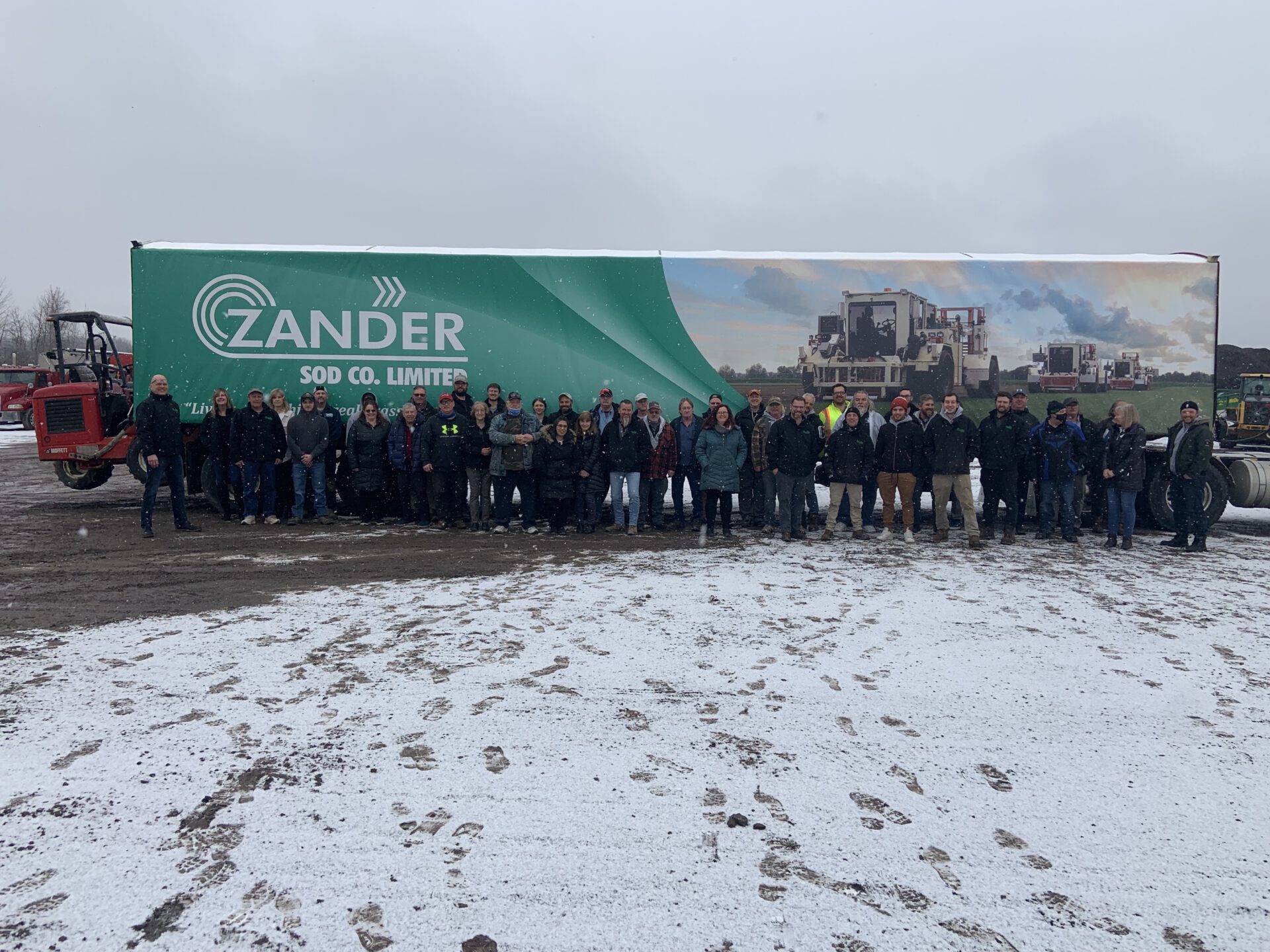 A group of people stands in front of a large trailer with "ZANDER SOD CO. LIMITED" branding, with snow on the ground and cloudy skies overhead.