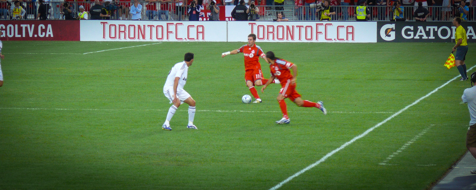 Two soccer players in red and white uniforms in action during a match, with a referee and spectators in the background, at Toronto FC's stadium.