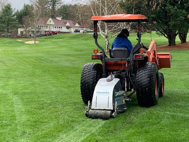 A person is operating a red tractor with a white Imants aerator attachment on a green golf course, near buildings under a cloudy sky.
